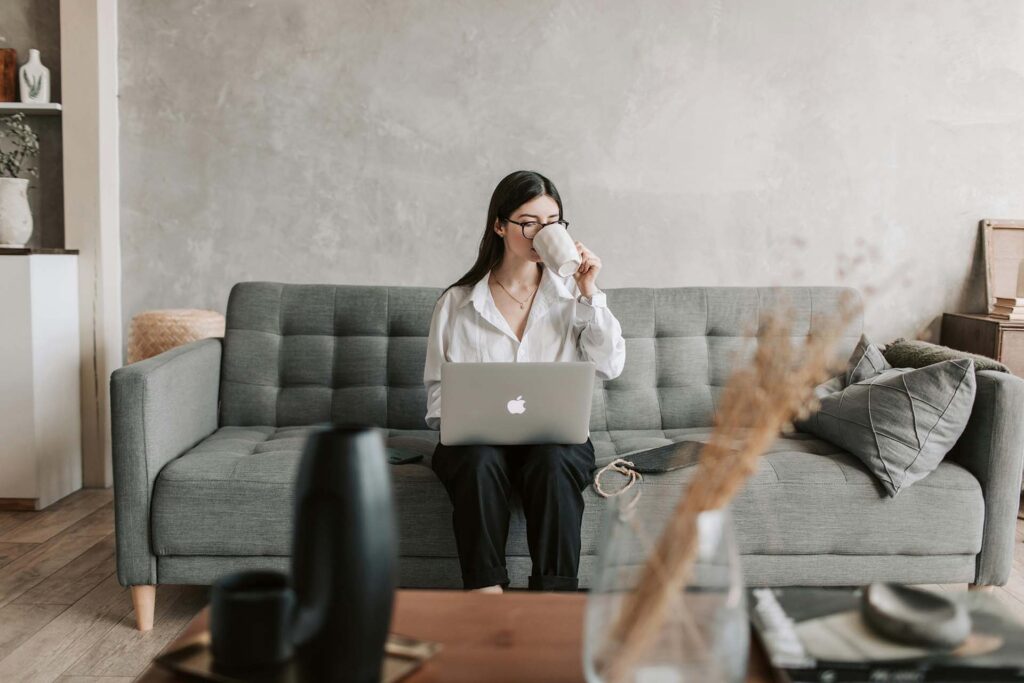 A woman sitting on a sofa, drinking coffee, and typing on a laptop 